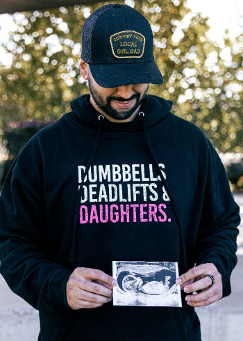 Man wearing a black hoodie with text, holding a ultrasound photo outdoors.